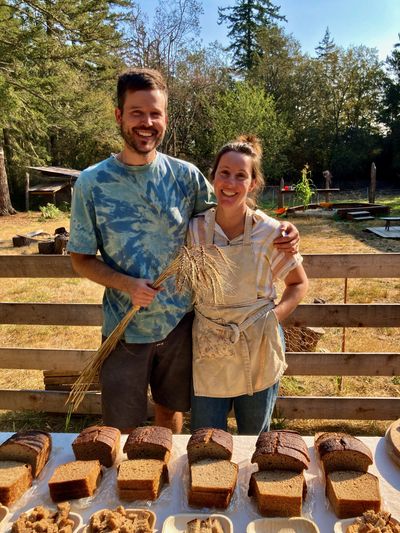 Nathan Hodges holding wheat stalks beside Sage Dilts, in front of her beautiful bread. Both on their farm on Lopez Island.