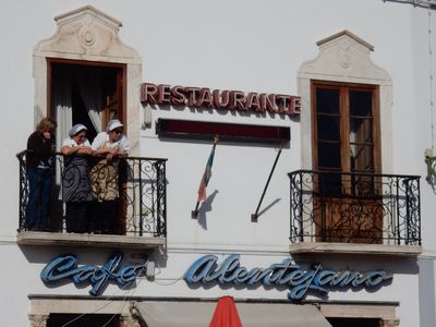 three women, dressed in aprons, watch the truck parade from a balcony above a restaurant.