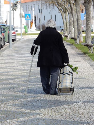An elderly lady, walking with a crutch-cane in her left arm, pulling a basket full of groceries with her right arm.